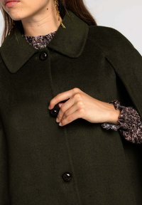 Cape en laine vert foncé avec un col arrondi, ornée de boutons noirs. Le modèle porte également un chemisier texturé et à motifs avec des poignets à volants.