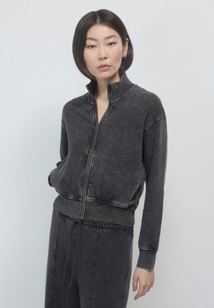 Schwarze Zip-Up-Jacke mit hohem Kragen. Hergestellt aus einem weichen, strukturierten Stoff mit gerippten Bündchen und Saum sowie sichtbaren Nähten.