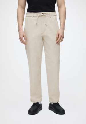 Hombre vistiendo pantalones beige con cordón, zapatos negros y una camiseta negra sin mangas, de pie frente a un fondo claro y simple.