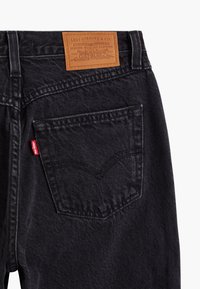 Jean en denim noir avec une étiquette en cuir, coutures visibles et un design standard pour la poche arrière. La texture semble durable et lisse.