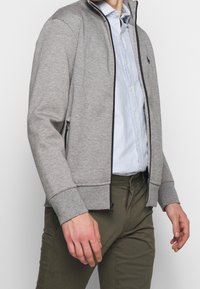 Grå jacka med dragkedja, tillverkad av mjukt tyg, med hög krage, sidofickor med dragkedjor och kontrasterande svarta detaljer. Enkeldesign.