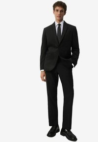 Musta puku ruudullisella kuviolla, istuva malli, teräväkaulukset, valkoinen paita ja musta solmio. Mustat kengät täydentävät muodollista ilmettä.