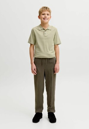 Garçon blond debout, portant un polo vert clair à manches courtes, un pantalon marron et des chaussures noires sur un fond clair uni.