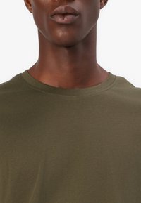 Tricou de culoare verde măsliniu cu un finisaj neted și texturat. Prezintă un guler rotund clasic și mâneci scurte. Design simplu și minimalist.