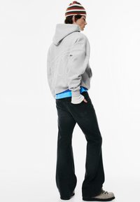 Grijze hoodie met capuchon en voorzak, blauwe blouse eronder, zwarte uitlopende spijkerbroek en een meerkleurige gestreepte muts met texturen en patronen.