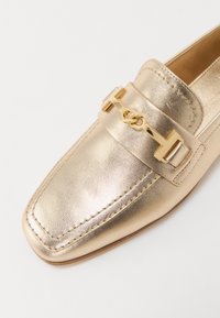 Mocassin en cuir doré avec un bout carré, présentant un accent décoratif en métal doré sur la sangle et des coutures ton sur ton le long des bords.