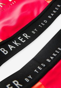 Punainen ja oranssi kiiltävä kangas mustalla jousto-ominaisuudella. Nauhassa on keskellä valkoisella tekstillä "TED BAKER".