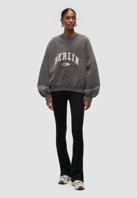 Sudadera gris de gran tamaño con el texto "BERLIN", cuello de canalé y mangas anchas, combinada con pantalones ajustados negros y zapatillas de deporte beige.