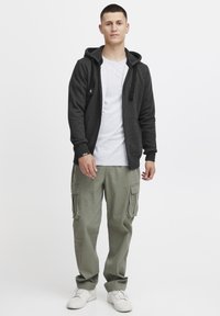 Sudadera gris con cremallera y cordones, sobre una camiseta blanca. Combinada con pantalones cargo verdes y zapatillas blancas. Fondo simple.