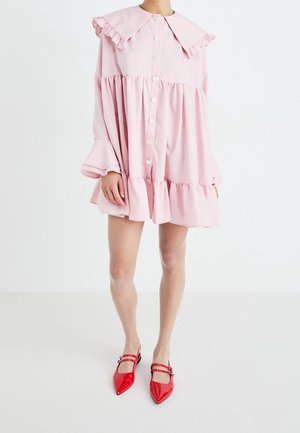 Osoba ubrana w luźną, jasnoróżową sukienkę z falbankami przy kołnierzu i brzegu, zapinaną na guziki, oraz czerwone baleriny z podwójnymi paskami i spiczastym czubkiem.