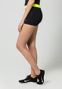 Svarta atletiska shorts med ett neongult midjeband, som har en åtsittande design och slät textur. Bärs med svarta löparskor.