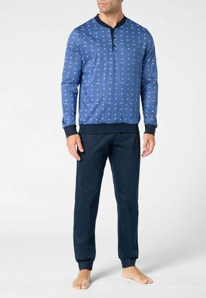 Uomo che indossa una maglia a maniche lunghe blu con motivo, polsini e colletto neri, abbinata a pantaloni blu scuro semplici, in piedi a piedi nudi.