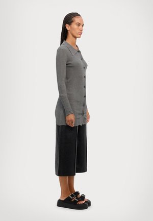 Cardigan grigio a coste con colletto e bottoni, abbinato a pantaloni neri larghi e cropped e sandali neri con doppia fibbia.