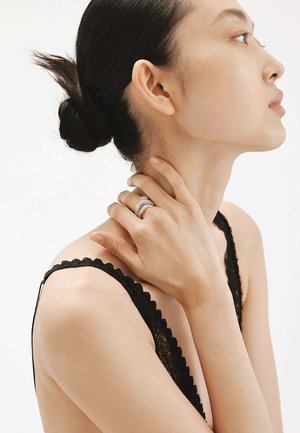 Anillo de plata con diseño entrelazado, llevado en una mano de piel clara, sujetando el cuello. El modelo lleva un top sin mangas de encaje negro con bordes festoneados.