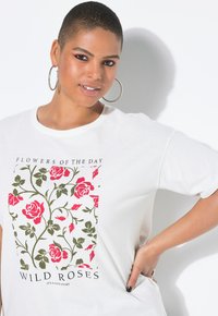 Femme aux cheveux courts et boucles d'oreilles en forme de créole portant un T-shirt blanc avec un imprimé de rose rouge et de feuille verte et le texte "Roses Sauvages."