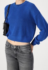 Maglione corto lavorato a maglia blu con scollatura rotonda e orli a coste, abbinato a jeans grigi a vita alta con dettagli di bottoni e tasche.
