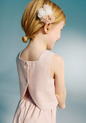 Jeune fille aux cheveux blonds attachés en un nouage bas, portant une robe rose sans manches avec des boutons dans le dos et une barrette fleurie dans les cheveux.