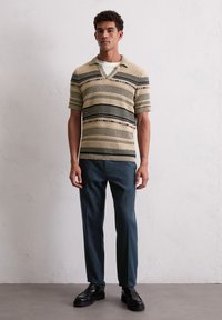 Beżowy golfowy top z czarnymi poziomymi paskami, zestawiony z ciemnoniebieskimi spodniami i czarnymi butami. Teksturowana tkanina z kołnierzykiem.