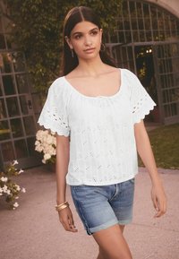 Haut en coton blanc avec des manches échancrées, des détails en œillets et un col rond. Porté avec un short en denim et plusieurs bracelets en or.