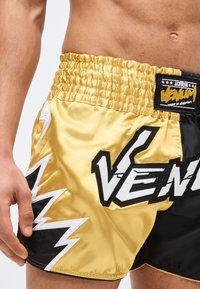 Κοντινό πλάνο ατόμου που φοράει χρυσά και μαύρα αθλητικά σορτς Venum με λεπτομέρεια αστραπής, εμφανίζοντας γυμνό κορμό και χαλαρό αριστερό χέρι.