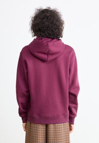 Sudadera con capucha de color marrón burdeos y corte relajado, con puños y dobladillo de canalé. Textura suave, sin patrones visibles, llevada sobre una falda de cuadros en tonos marrones.