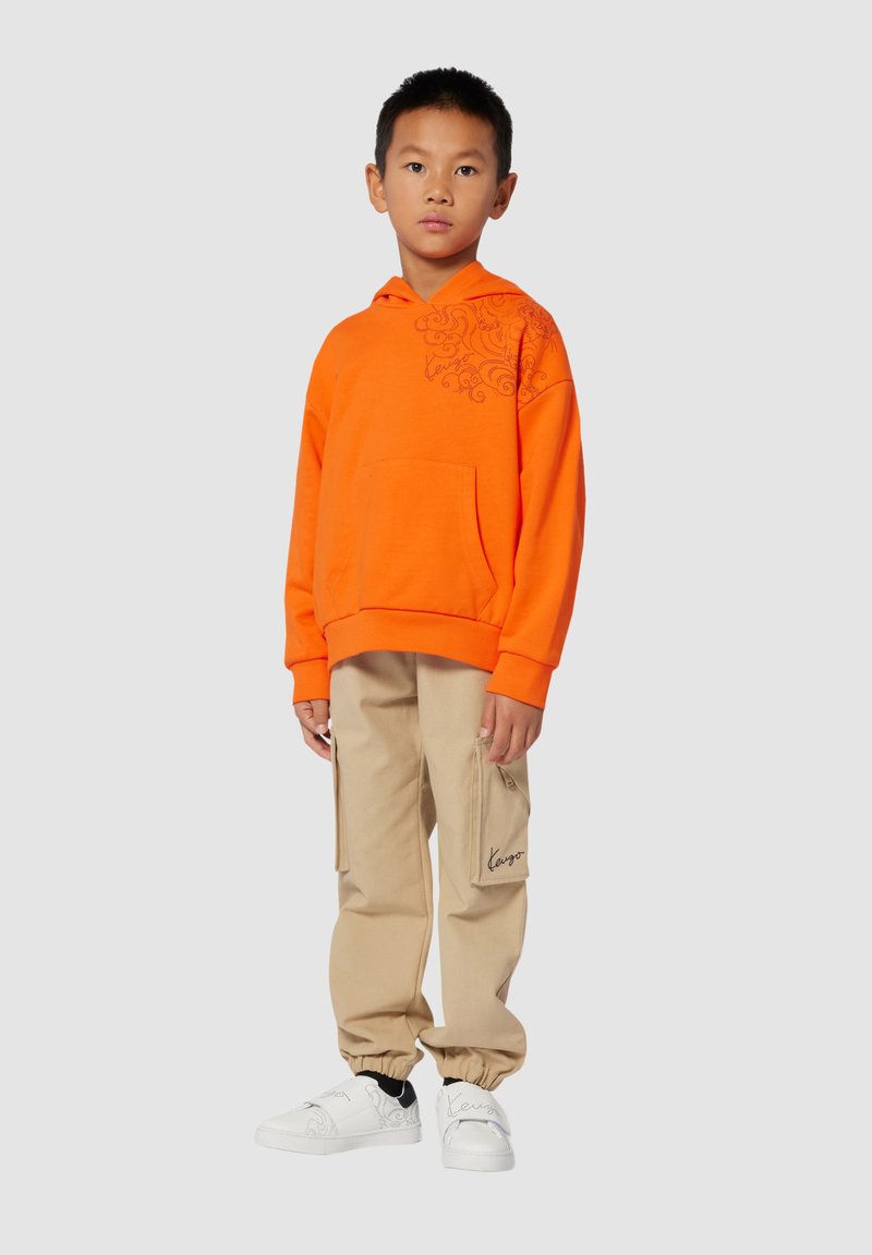 Een oranje hoodie met een voorzak en decoratieve stiksels, gecombineerd met beige cargobroeken met zijzakken en witte sneakers.
