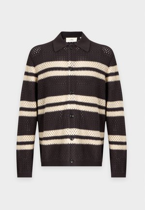 Jeune homme portant un cardigan rayé marron et beige, un jean gris délavé, une casquette beige, des chaussures beiges et portant un sac à bandoulière beige et marron.