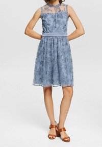 Vestido azul claro con bordado floral, cuello transparente, cintura ajustada y falda acampanada; combinado con sandalias de tacón con tiras marrones.