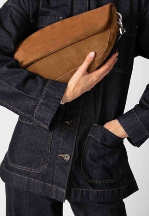 Braune Wildleder-Clutch, die vor einer dunklen Jeansjacke gehalten wird, mit glatter Textur und silbernem Reißverschluss-Akzent.