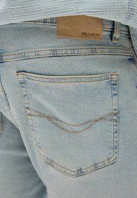Světlé modré džíny z denimu s viditelnou zadní kapsou a zakřiveným švem. Hnědá kožená nášivka v pase nese označení "PULL&BEAR."