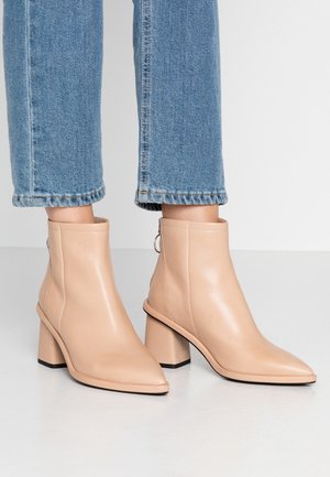 Bottines beiges à bout pointu avec talons blocs portées avec un jean court bleu sur fond gris.