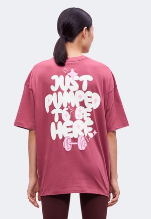Persoon met donker haar in een paardenstaart die een roze oversized T-shirt draagt met witte tekst "Just pumped to be here" en een roze haltergrafiek op de achterkant.