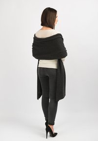 Mørkegrå wrapping lavet af blødt, tekstureret stof, der er draperet over en cremefarvet bluse. Sorte høje hæle og slim-fit mørke jeans fuldender outfittet.