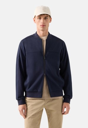 Ung mand iført en marineblå jakke med lynlås, beige bukser, tan t-shirt og en lys beige baseballkasket mod en ensfarvet baggrund.