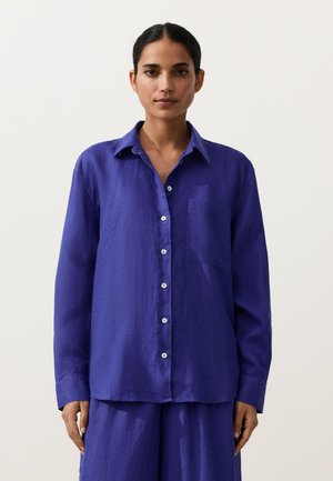 Žena s tmavými vlasmi, ktorá má na sebe voľnú fialovú košeľu s dlhými rukávmi a gombíkmi a zodpovedajúce nohavice, stojaca pred jednoduchým pozadím.