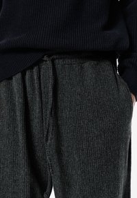 Közelkép egy sötét bordázott kötött nadrágot viselő emberről, amelynek zsinórja van, és egy sötétkék bordázott pulóvert visel. Egyik kéz a zsebében van.