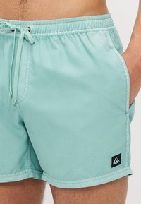 Calções de banho azul claro feitos de tecido suave. Apresentam uma cintura elástica com cordão, bolsos laterais e uma pequena etiqueta com o logótipo.