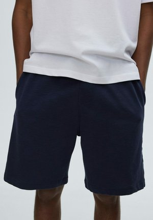 Persona con pantalones cortos azul oscuro y una camiseta blanca con las manos en los bolsillos contra un fondo claro y liso.