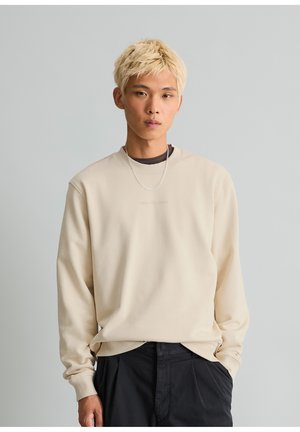 Jeune homme aux cheveux blonds courts, portant un sweat-shirt beige, un pantalon noir et une chaîne en argent autour du cou, debout sur un fond uni gris clair.