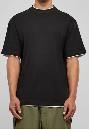 Mand iført en løs sort kortærmet t-shirt med hvid kant ved krave og ærmer, kombineret med olivengrønne bukser.