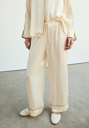 Persona con pantalones de satén sueltos de color crema con cintura elástica y zapatillas de satén a juego sobre un suelo de cemento.