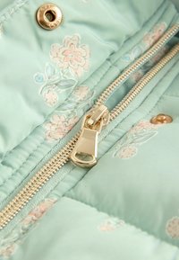 Hellblau, gepolsterte Jackenstoff mit floralem Druck; verfügt über einen goldenen Reißverschluss und Druckknopfdetails. Glatte Textur mit gesteppten Abschnitten.