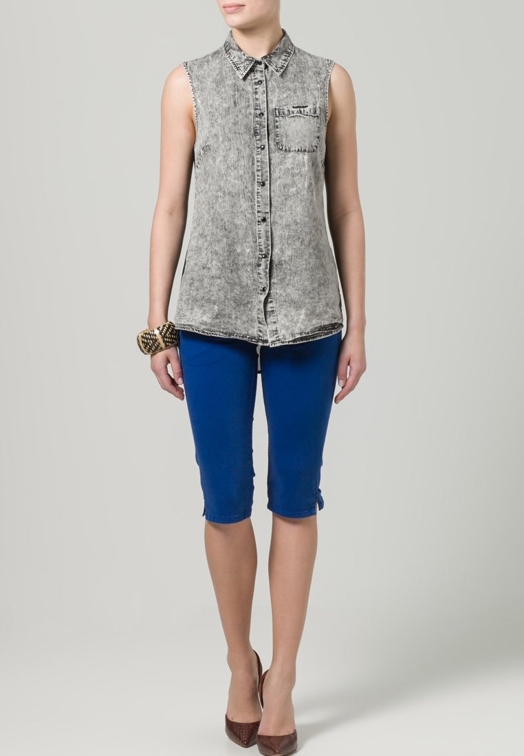 Canotta in denim grigio chiaro con taschino frontale, abbinata a pantaloni blu fino al ginocchio. Tacchi marroni e un braccialetto decorativo completano l'outfit.