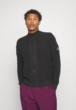 Maglione zip-up da uomo in texture a costine nera, con colletto alto e patch del logo CK sulla manica sinistra. Indossato con pantaloni viola.