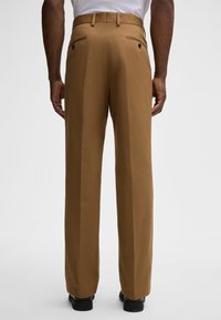 Pantaloni marroni su misura con gamba larga, caratterizzati da una texture liscia, cuciture doppie e due tasche posteriori con chiusura a bottone.