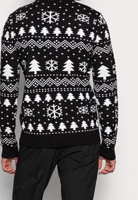 Person klädd i svart tröja med mönster av vita julgranar och snöflingor, sedd bakifrån med svarta byxor.