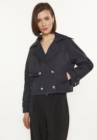 Giacca nera accorciata con fronte doppiopetto, ampio colletto, bottoni metallici e tasche laterali. Tessuto liscio, vestibilità sartoriale.
