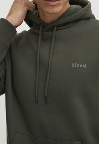 Hoodie verde oliva realizzato in tessuto morbido, dotato di tasca a marsupio, cordini e piccolo logo "blend" ricamato sul petto.