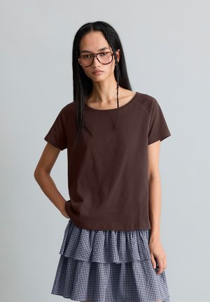 Femme aux longs cheveux noirs et lunettes, portant une chemise marron à manches courtes et une jupe à couches bleue et blanche à carreaux, debout devant un fond gris.