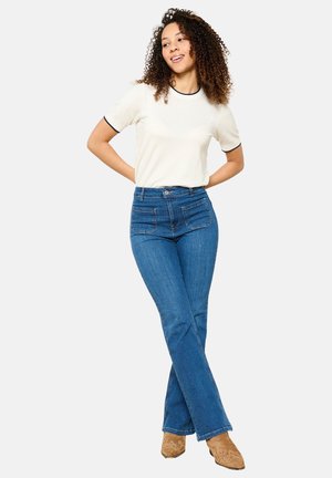 Camicetta bianca a maniche corte con bordi neri, jeans blu a vita alta svasati con tasche anteriori e stivaletti con tacco color beige.